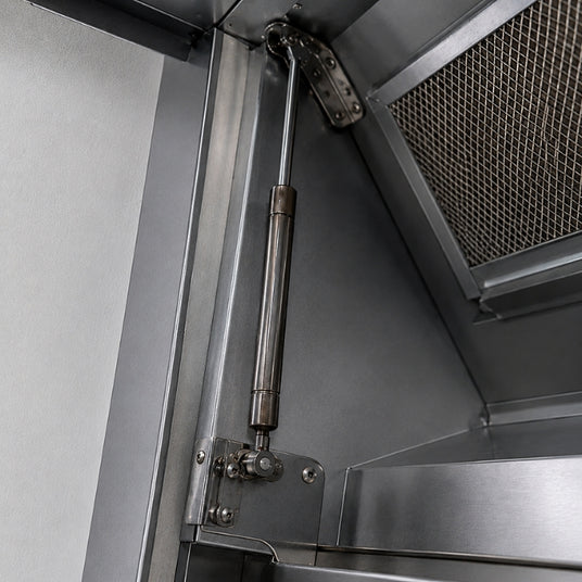 Detailaufnahme einer geöffneten Kopffreihaube mit scharf sichtbarem seitlichem Gasdruckdämpfer, Metallhalterung und Fettfilter im Hintergrund.